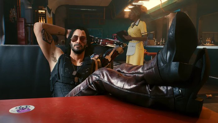 Screenshot z Cyberpunka 2077. Postać Johnny'ego Silverhanda siedzi w bufecie z nogami położonymi na stole naprzeciwko gracza.