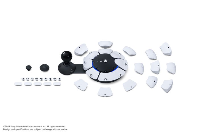 Obrazek to marketingowe zdjęcie przedstawiające kontroler PlayStation Access na białym tle. Pad jest płaski okrągły, z lewej strony ma doczepianą gałkę. Na potrzeby zdjęcia pokazano wszystkie przyciski, jakie można zamontować w urządzeniu.
