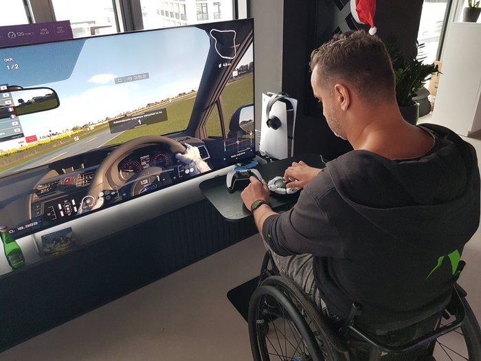 Zdjęcie przedstawia mężczyznę na wózku w czarnym ubraniu, który gra w Gran Turismo za pomocą PlayStation Access. Po lewej stronie widoczny jest duży telewizor z uruchomioną grą.
